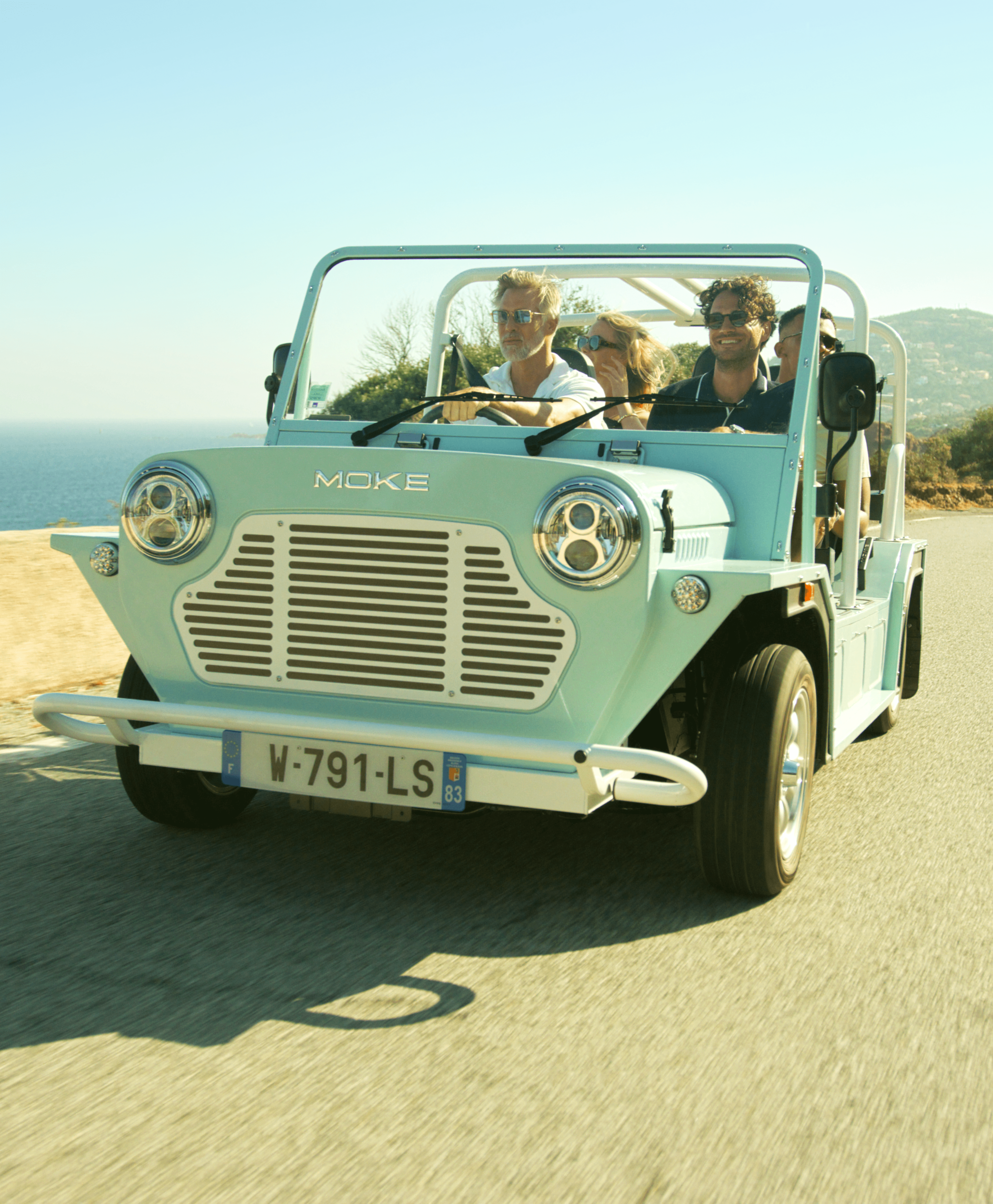Group of friends driving in a Moke vehicle along the side of the beach on a sunny day.