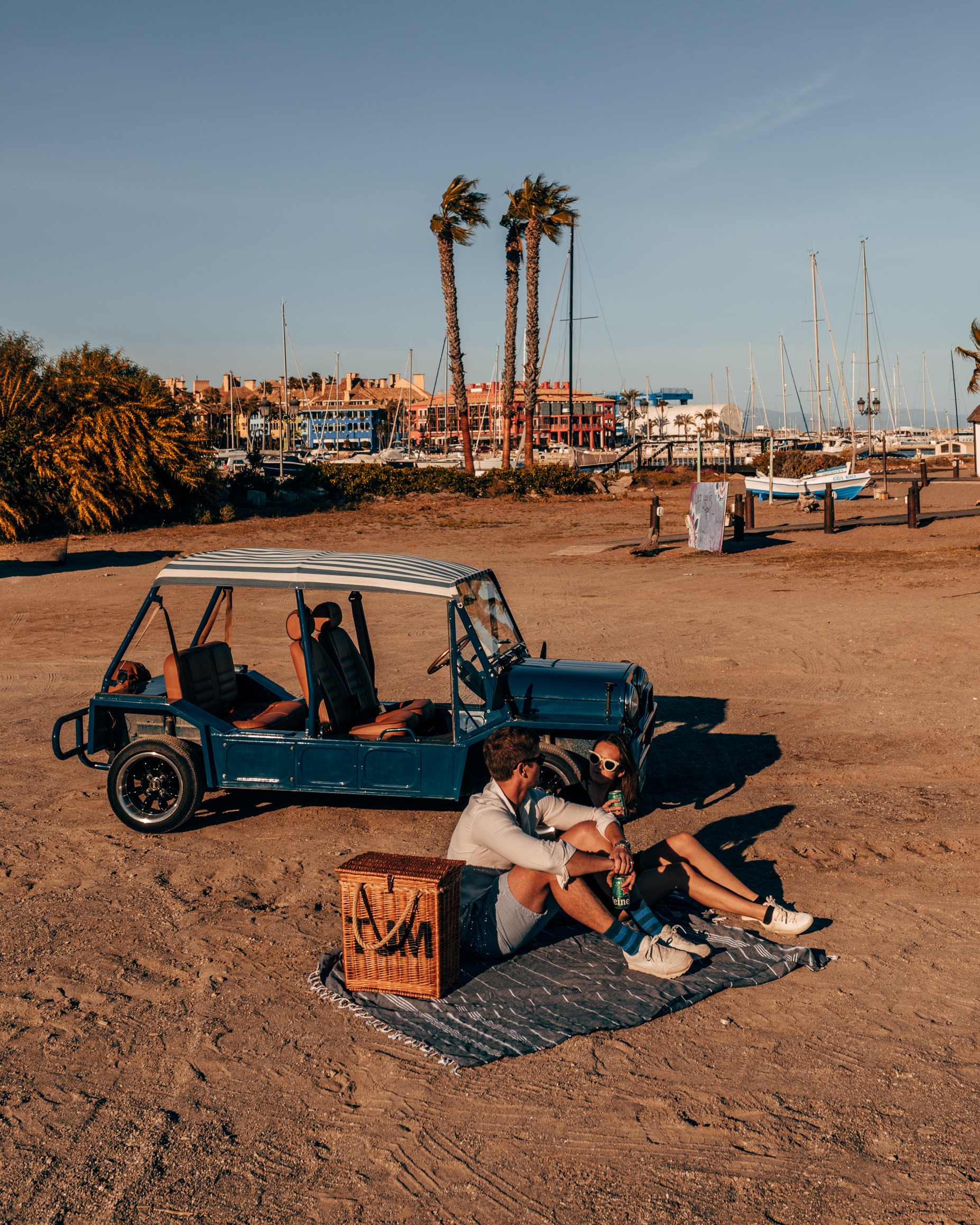 Two people enjoying a beach picnic next to a Moke in Marbella, Spain, during a beautiful sunset.