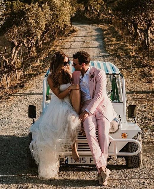 Newlywed couple sitting on the front of a Moke vehicle, looking at each other affectionately, parked in the middle of a rocky road on a sunny day.