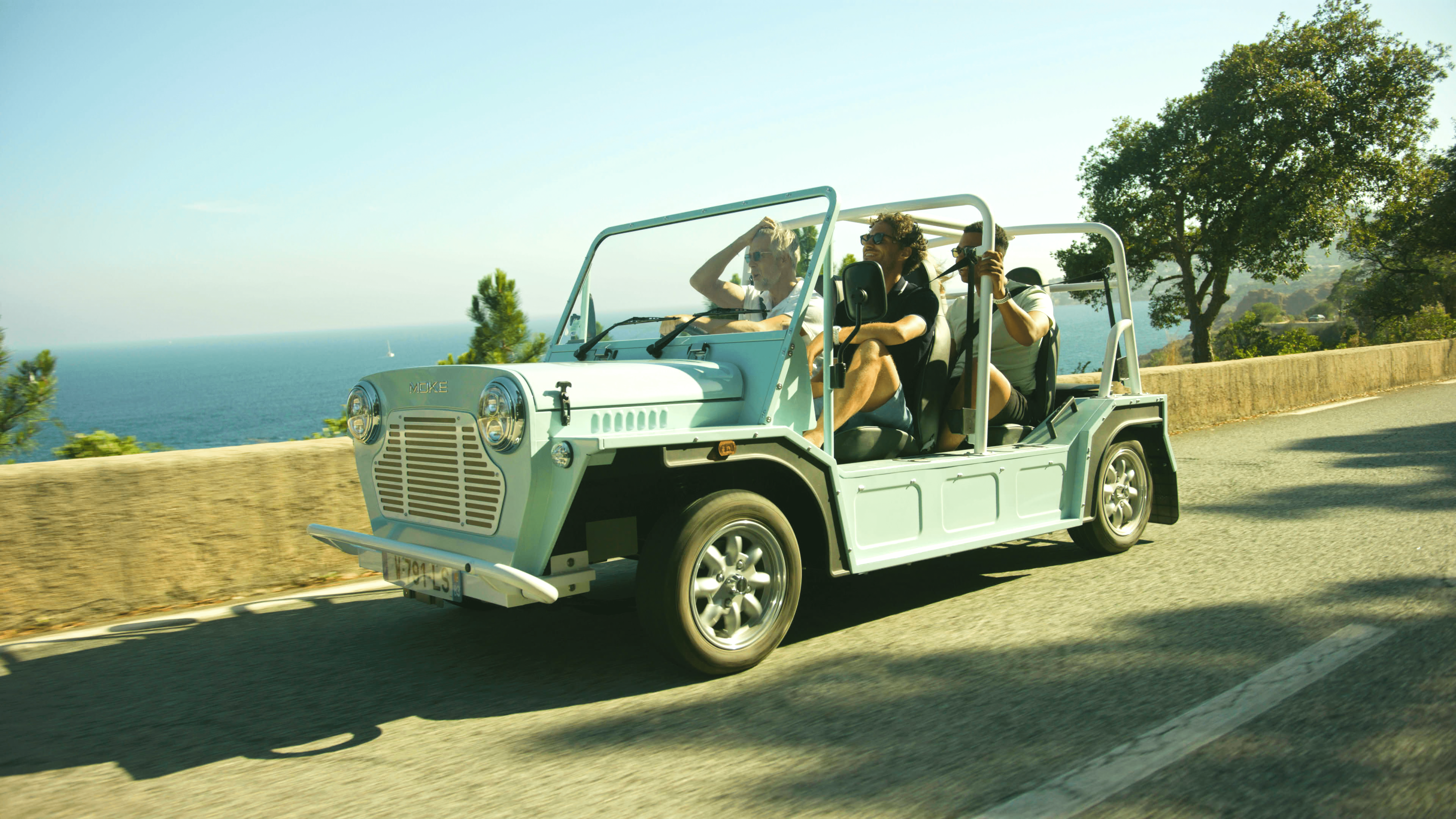 Group of friends enjoying a road trip in a Moke vehicle with the sea in the background on a sunny day.
