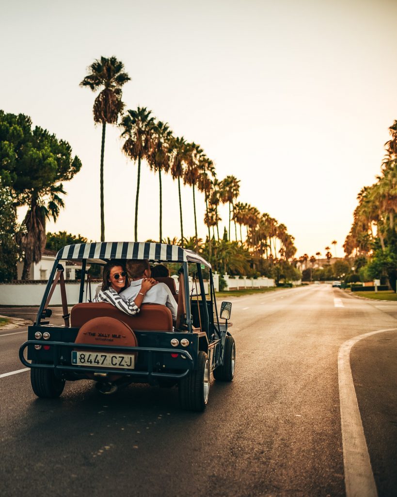 Group of close friends driving a Moke along a palm tree-lined road on a sunny afternoon.